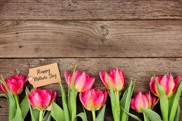 Bottom border of pink flowers with Mothers Day gift tag against a rustic wood background - Foto, Imagen