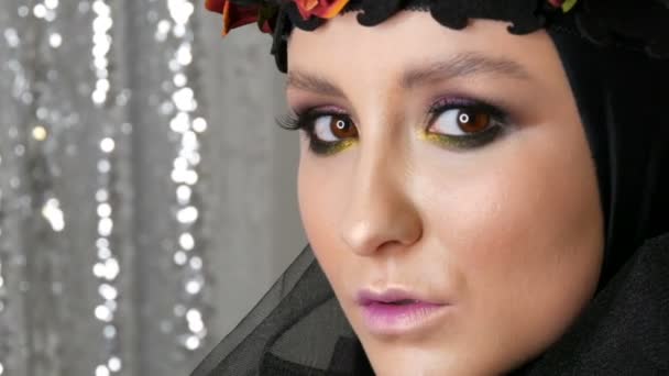 Profi-Model mit wunderschönem Make-up posiert mit schwarzer Mütze und Kranz auf dem Kopf vor der Kamera auf silbernem Hintergrund im Bild einer schwarzen Witwe. Mode im Trend - Filmmaterial, Video