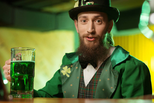 Dark-haired bearded young man in a green costume with a shamrock symbol sitting at the table - Photo, Image