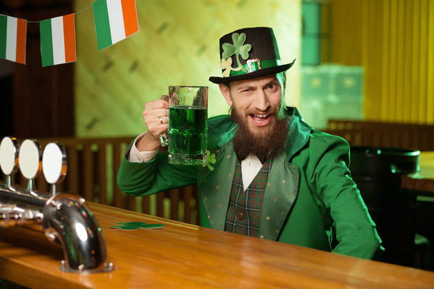 Dark-haired bearded young man in a leprechaun hat with shamrock sitting at the irish pub - Photo, Image