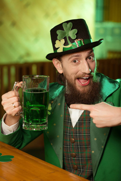 Dark-haired bearded young man in a leprechaun hat with shamrock pointing to the glass with beer - Fotó, kép