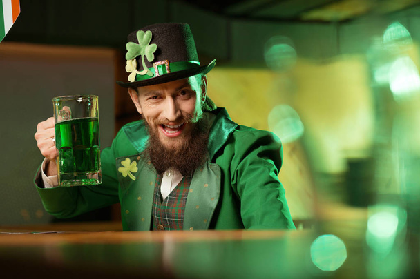 Dark-haired bearded young man in a leprechaun hat with shamrock looking funny - Foto, Imagen