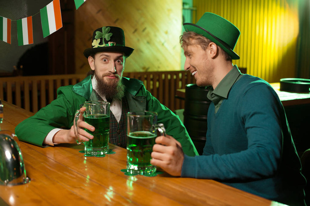 Dark-haired bearded young man in a leprechaun hat and his friend having great time in the pub - Foto, afbeelding