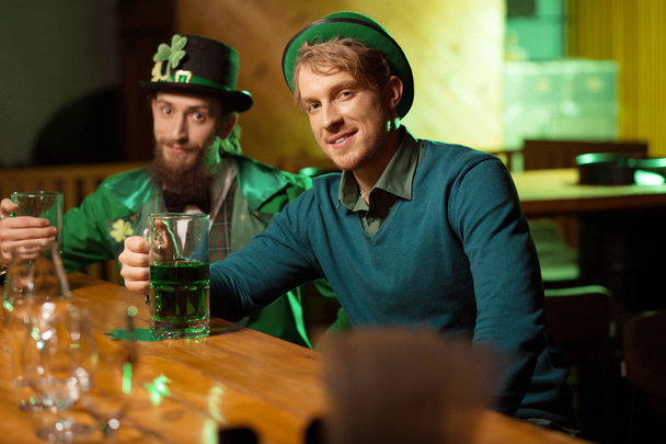 Fair-haired young handsome man in a green hat and his friend in a leprechaun costume sitting at the bar counter - Foto, Imagen