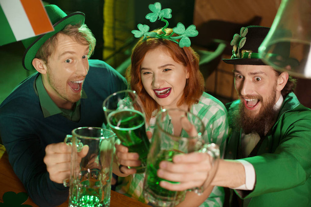 Red-haired pretty girl in a green checkered shirt and men in st.patricks day costumes raising glasses with green beer - Фото, изображение