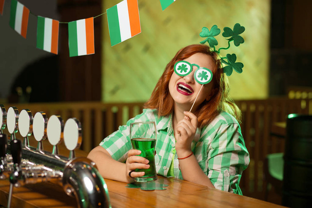 Red-haired pretty girl in a green checkered shirt having fun with eyewear with shamrock symbols - Photo, Image