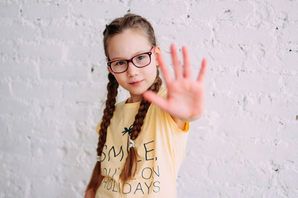 Serious long hair tweens girl in glasses pulling hand towards camera on white wall background - Photo, Image