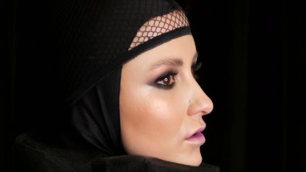 Επαγγελματικό μοντέλο κορίτσι με όμορφο μακιγιάζ ποζάρει σε ένα μαύρο καπάκι στο κεφάλι της μπροστά από την κάμερα σε μαύρο φόντο στην εικόνα μιας μαύρης χήρας. Υψηλής μόδας - Πλάνα, βίντεο