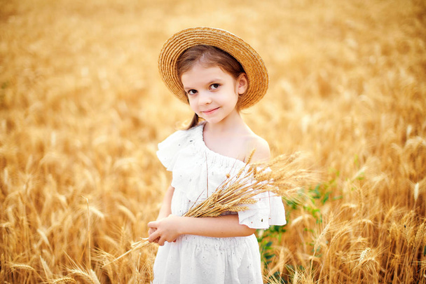 Happy child in autumn wheat field. Beautiful girl in white dress and straw hat have fun with playing, harvesting - Foto, Bild