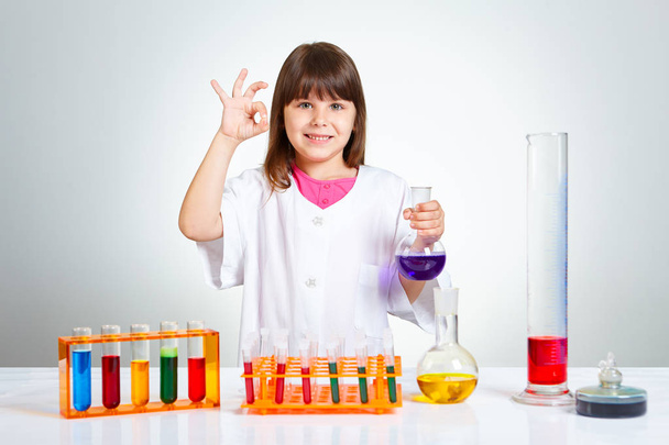junges 6-jähriges Mädchen gestikuliert großartig mit der rechten Hand, während sie in der anderen Hand das Reagenzglas mit lila Flüssigkeit hält  - Foto, Bild
