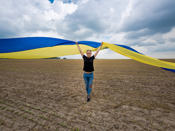 Νεαρό εκπληκτικό όμορφο κορίτσι με μια τεράστια ουκρανική σημαία στο πεδίο. Η ημέρα της ανεξαρτησίας της Ουκρανίας. - Φωτογραφία, εικόνα