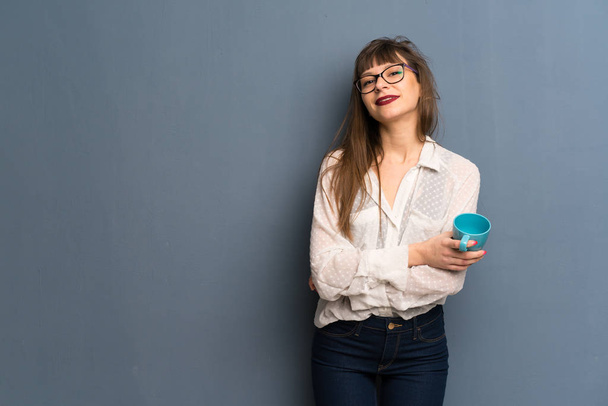Γυναίκα με τα γυαλιά πάνω από μπλε τοίχο, κρατώντας ένα ζεστό φλιτζάνι καφέ - Φωτογραφία, εικόνα