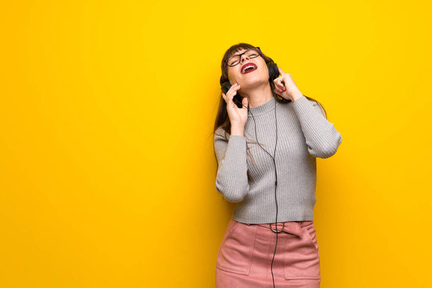 Γυναίκα με τα γυαλιά πάνω από κίτρινο τοίχο, να ακούτε μουσική με ακουστικά - Φωτογραφία, εικόνα