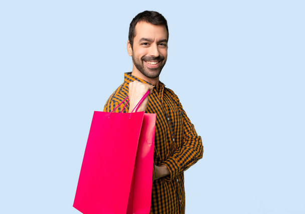 Hombre con bolsas de compras manteniendo los brazos cruzados en posición lateral mientras sonríe sobre un fondo azul aislado
 - Foto, Imagen