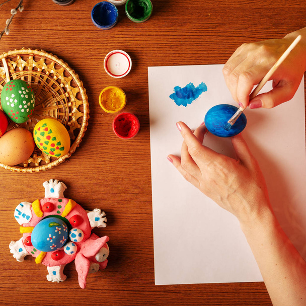 De kunstenaar in glazen voert ontwerpwerk. Schildert eieren met verf. Lente, warme sfeer. Op de tafel zijn de takken van wilg en Lila met groene bloei bladeren. Veelkleurige Pasen stilleven - Foto, afbeelding