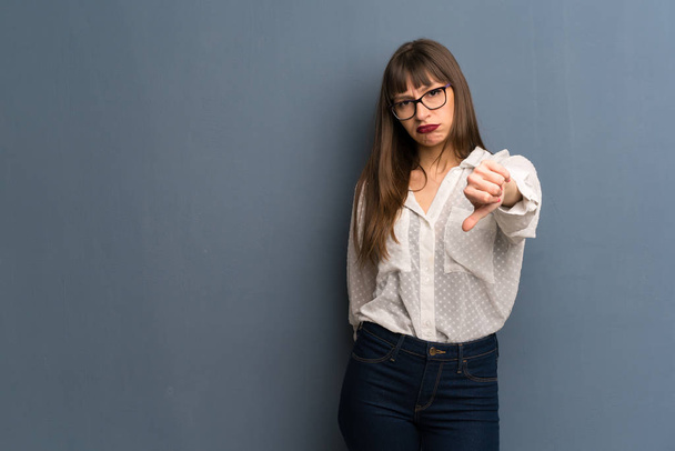 Mujer con gafas sobre pared azul mostrando el pulgar hacia abajo signo con expresión negativa
 - Foto, Imagen