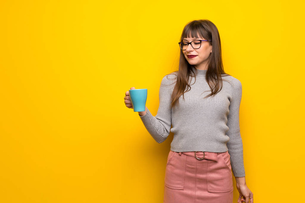 Γυναίκα με τα γυαλιά πάνω από κίτρινο τοίχο, κρατώντας ένα ζεστό φλιτζάνι καφέ - Φωτογραφία, εικόνα