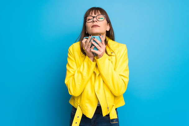 Молодая женщина в жёлтой куртке на синем фоне держит чашку горячего кофе
 - Фото, изображение