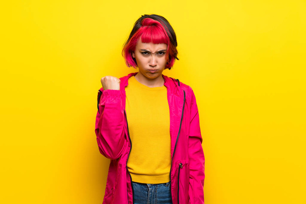 Jeune femme aux cheveux roses sur un mur jaune avec un geste de colère
 - Photo, image