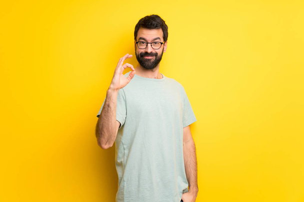 Homem com barba e camisa verde mostrando um sinal ok com os dedos
 - Foto, Imagem