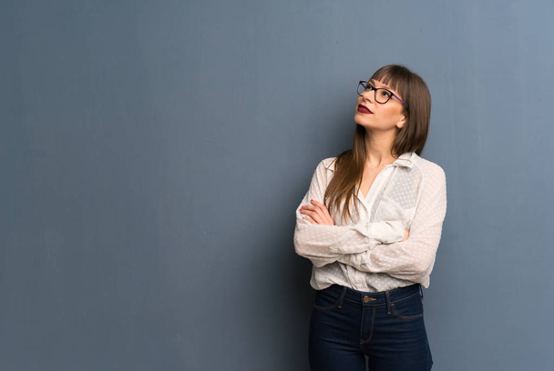 Γυναίκα με γυαλιά πάνω από τον μπλε τοίχο κοιτάζοντας ψηλά, ενώ χαμογελάει - Φωτογραφία, εικόνα