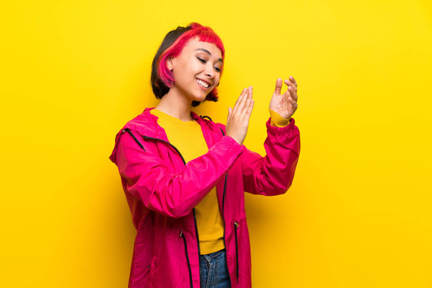 Nuori nainen, jolla on vaaleanpunainen tukka keltaisen seinän päällä, taputtaa esityksen jälkeen konferenssissa
 - Valokuva, kuva