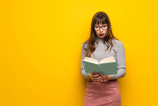 Mulher com óculos sobre parede amarela surpreendida enquanto aprecia a leitura de um livro
 - Foto, Imagem
