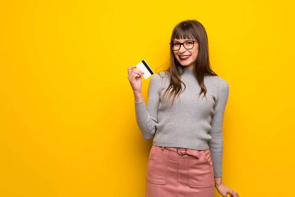 Жінка в окулярах над жовтою стіною тримає кредитну картку
 - Фото, зображення