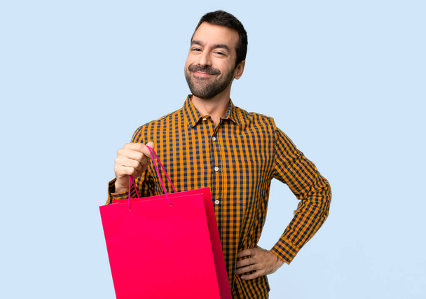 Homem com sacos de compras posando com os braços no quadril e sorrindo no fundo azul isolado
 - Foto, Imagem