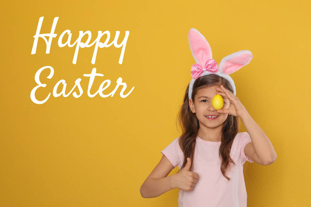 Маленькая девочка в бандаже в ушах кролика держит пасхальное яйцо возле глаз на цветном фоне. Счастливая Пасха
 - Фото, изображение