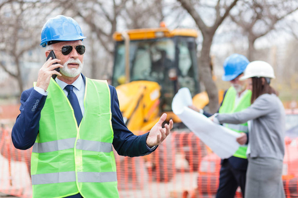 Súlyos vezető építész vagy beszél a telefonon, miközben egy építkezésen dolgozó üzletember - Fotó, kép