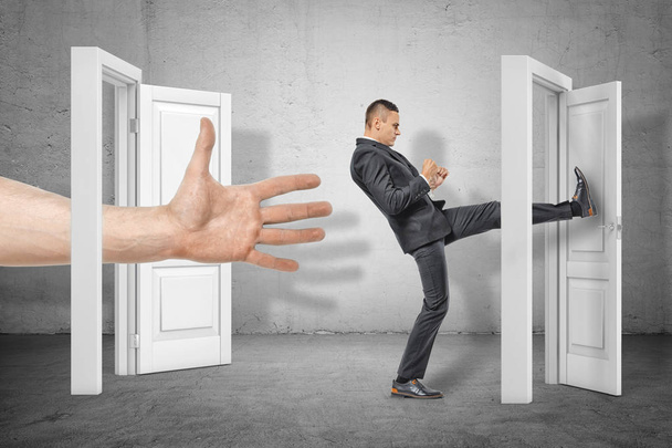 Businessman kicking a door and big hand appearing out of an open door on grey wall background - Foto, afbeelding