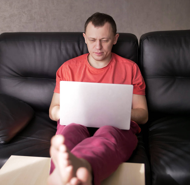 Νεαρός ελεύθερος επαγγελματίας εργάζεται στο σπίτι σε ένα φορητό υπολογιστή, ενώ κάθεται στον καναπέ - Φωτογραφία, εικόνα