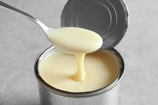 Condensed milk pouring from spoon into tin can on grey background, closeup. Dairy product - Fotografie, Obrázek