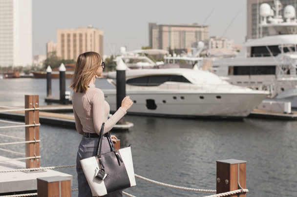 Young female model with long bob haircut, beige sweater, trousers and big shopper bag standing at pier or marina promenade and looking at yacht and boat - Zdjęcie, obraz