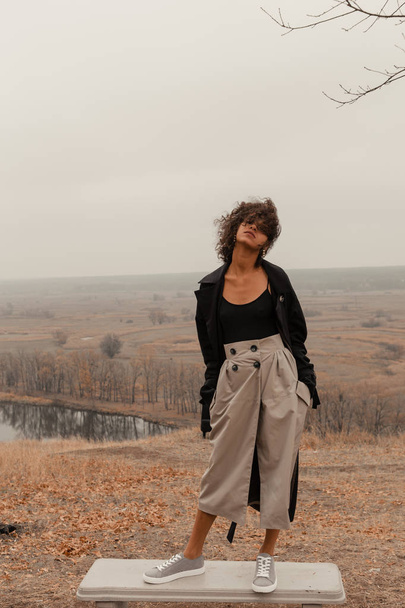 Beautiful girl with curly hair in a stylish suit walks against the backdrop of a beautiful landscape - Foto, Imagem