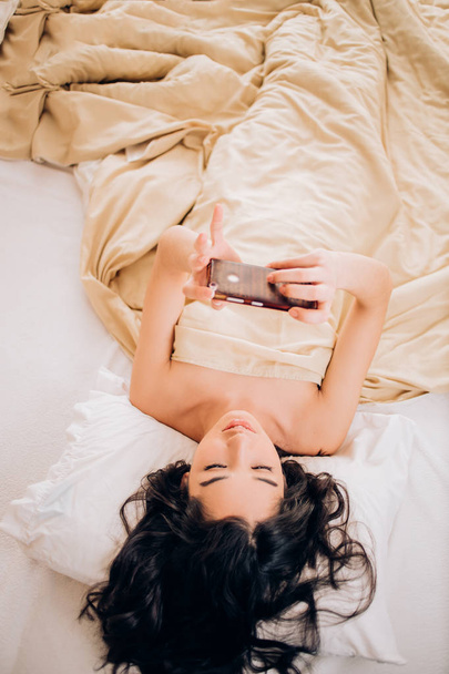 Beautiful young smiling brunette woman using phone in her bedroom - 写真・画像