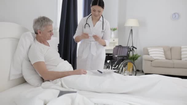 Asian nurse assists disabled senior man in bed - Materiał filmowy, wideo