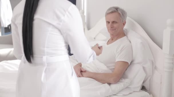 Charming doctor takes care of a disabled senior man in bed - Záběry, video