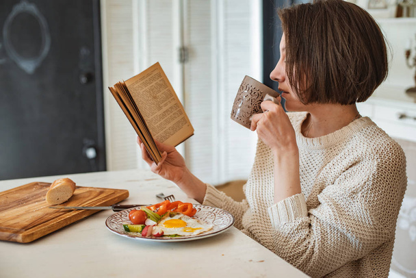 La joven está leyendo un viejo libro en el desayuno. Primer plano manos y mesa de la cena de ajuste
. - Foto, imagen