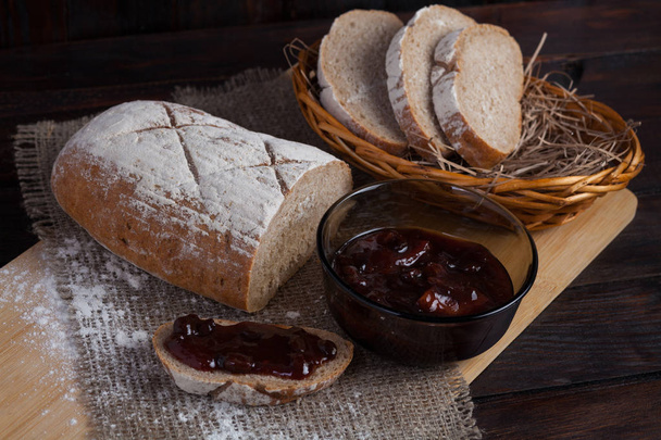 Pedaços recém fatiados pão em uma tigela de vime em um tabuleiro de carvalho com farinha espalhada em um fundo de madeira com tecido de linho ao lado de uma tigela de um delicioso geléia
 - Foto, Imagem