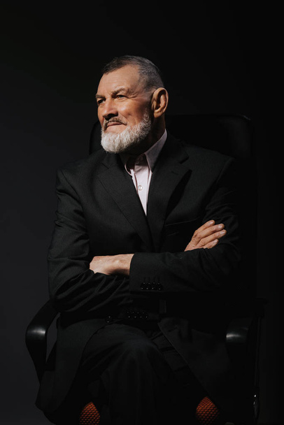 Portrait d'un bel homme barbu mature élégant sur un fauteuil avec les mains croisées. Coup de mode. Vêtements homme
  - Photo, image