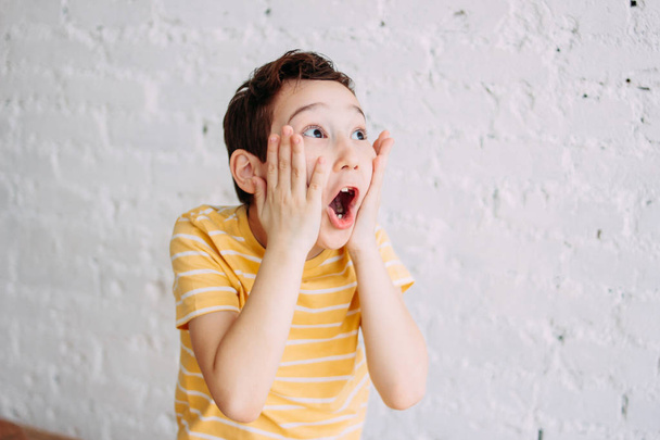 Cute tween surprised boy with funny face in yellow t-shirt isolated on white brick wall background - Photo, Image