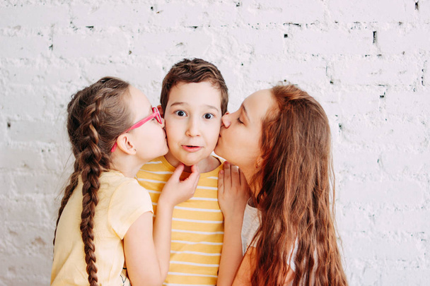 Girls sisters kissing boy brother with two sides isolated on white brick background - Photo, Image