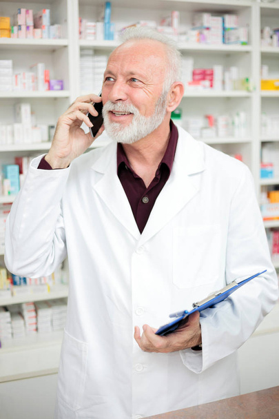 Older male pharmacist talking on the phone and holding a clipboard - Fotoğraf, Görsel