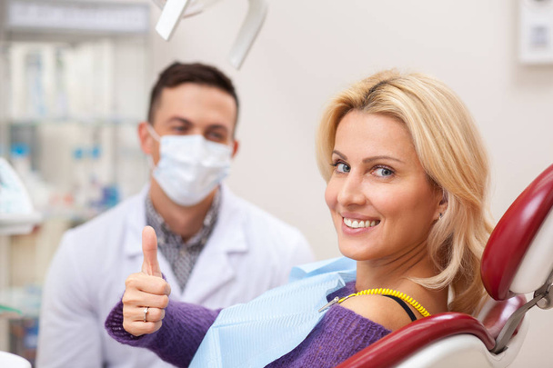 Gorgeous happy mature woman smiling to the camera, sitting in a dental chair at the clinic. Cheerful female patient showing thumbs up, her dentist on the background, copy space. Medical service - Zdjęcie, obraz