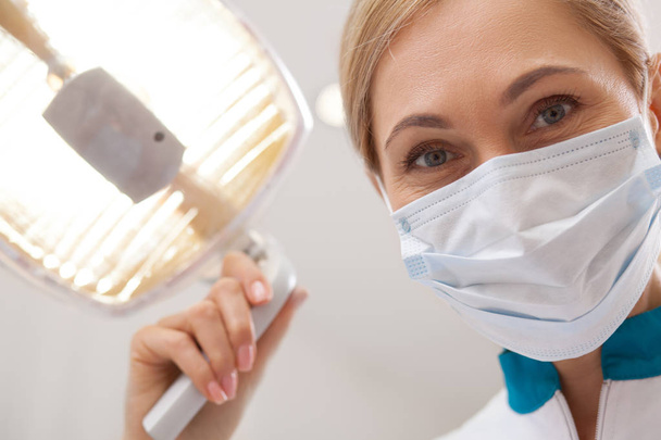 Nahaufnahme einer reifen Zahnärztin, die lächelt, medizinische Maske trägt und Zahnlampe justiert. professioneller Zahnarzt, der sich auf die Zahnuntersuchung vorbereitet und in die Kamera blickt. Der Patient muss sich gedulden. Medizinkonzept - Foto, Bild