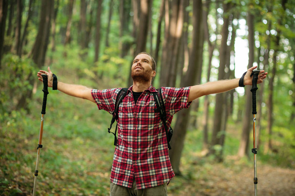 Elégedett fiatal férfi élvezi egy tökéletes békés pillanat alatt erdei kirándulás kockás inget. Arms kinyújtott és az égre néz - Fotó, kép