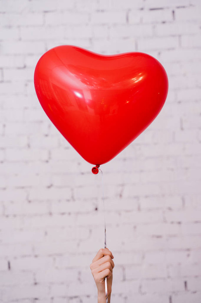 mano femenina sosteniendo globo de corazón rojo y posando sobre fondo de estudio de pared de ladrillo, concepto de fiesta de cumpleaños
  - Foto, Imagen