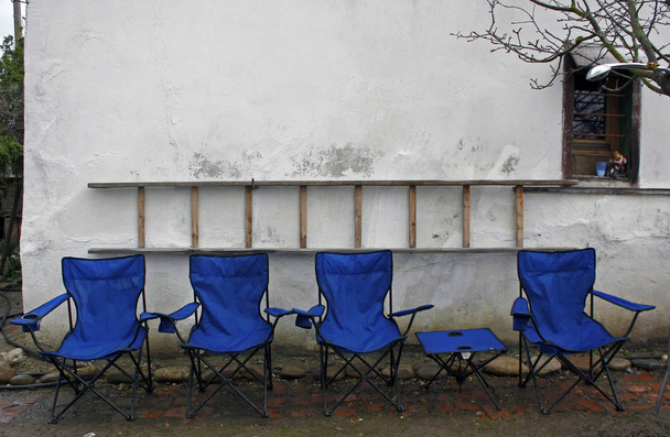 Empty blue folding camping chairs and table lined up in front of a rustic white wall with a ladder on the wall. - Photo, Image